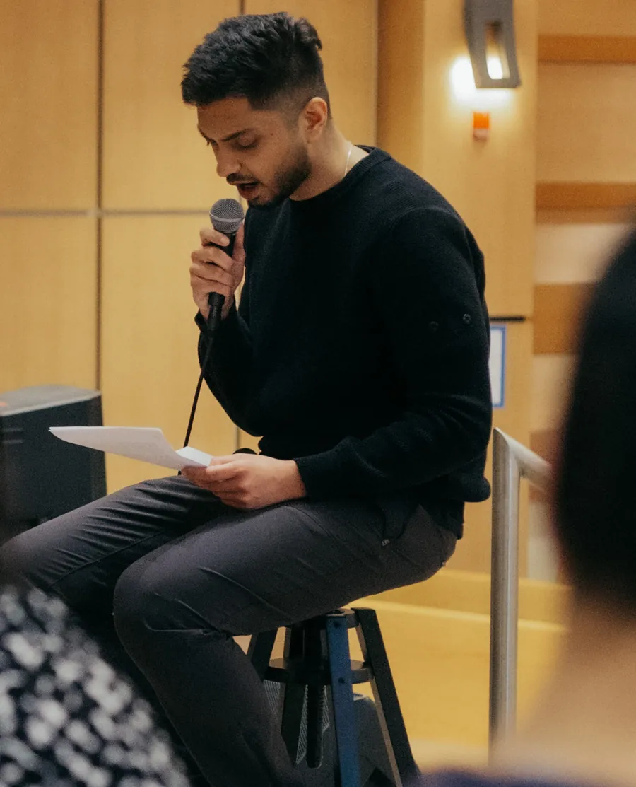 Shray Shrotriya seated on a stool holding a microphone in one hand and a sheet of paper in the other, mid-sentence, in a wood-panelled room. Members of the audience are blurred in the foreground.